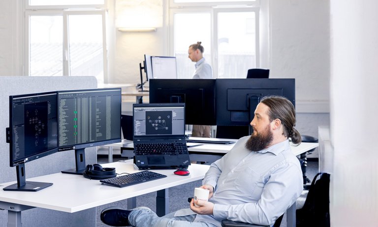 Photo of the TLK-Thermo office, a colleague sits relaxed at his desk during a coffee break. In the background you can see computer screens on which the MoBA automation is running.