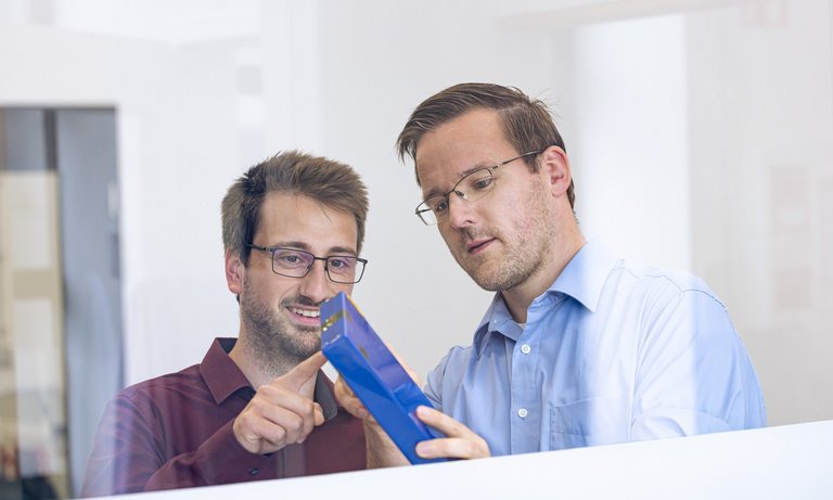 Foto auf dem ein Kollege einem anderen Kollegen etwas an einem blauen Batteriemodul zeigt, fotografiert durch eine Glasscheibe.