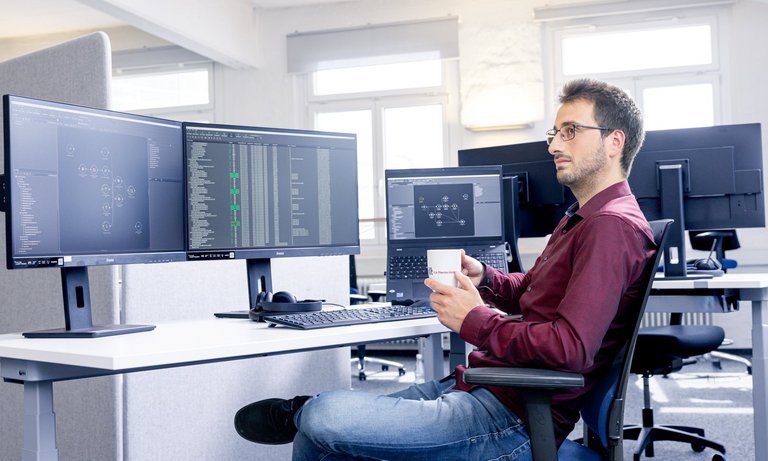 Photo of the TLK-Thermo office, a colleague sits relaxed at his desk during a coffee break. In the background you can see computer screens on which the MoBA automation is running.