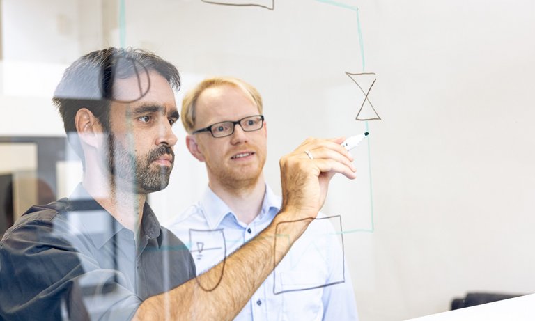 Photo of two colleagues standing behind a pane of glass and using it as a blackboard. A colleague draws and explains a refrigeration cycle.