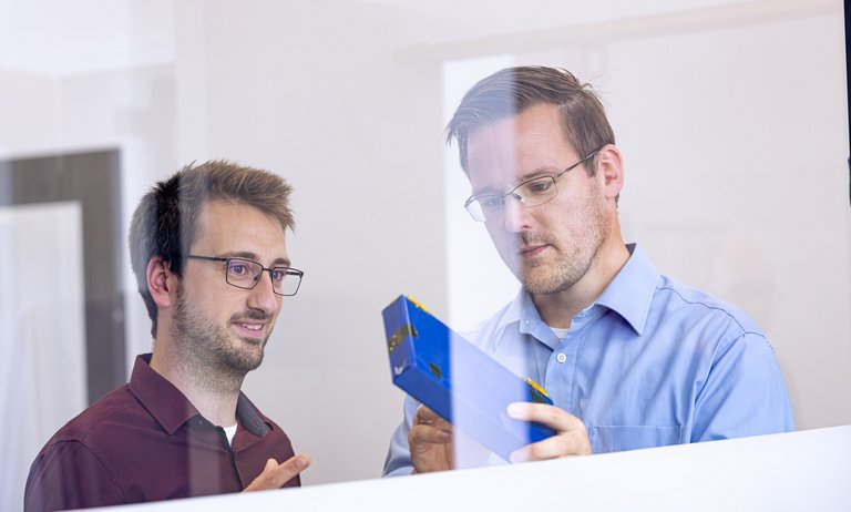 Foto, auf dem ein Kollege einem anderen Kollegen etwas an einem blauen Batteriemodul zeigt, fotografiert durch eine Glasscheibe.