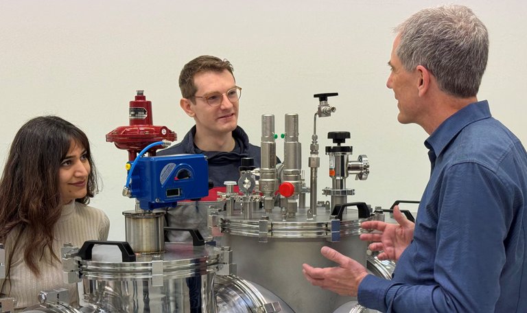 Photo of three employees standing around the installation of a small, decentralized H2 liquefaction plant. One colleague explains something about the test bench to the others.