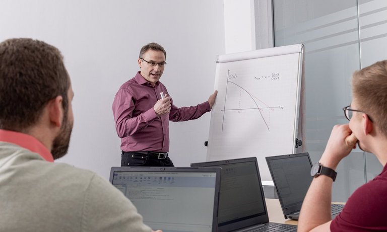 Foto einer Schulungssituation: Der Lehrer erklärt etwas an einer Papiertafel, drei Teilnehmer sitzen an Laptops und hören zu.